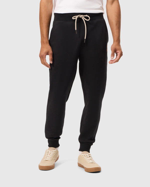 A person wearing a white shirt and Psycho Bunny's MENS JAMES PREMIUM SWEATPANT - B6P791D200, featuring black cotton-blend fabric and a beige drawstring, stands against a plain background. They also sport beige sneakers with brown soles.