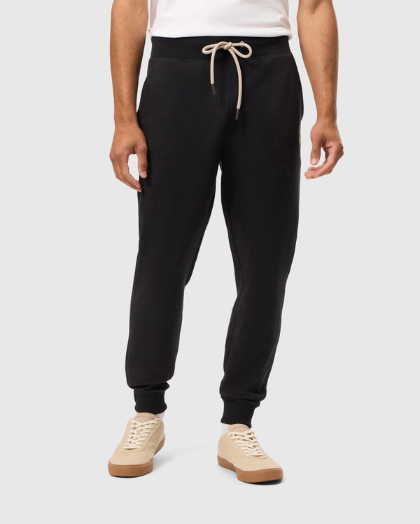 A person wearing a white shirt and Psycho Bunny's MENS JAMES PREMIUM SWEATPANT - B6P791D200, featuring black cotton-blend fabric and a beige drawstring, stands against a plain background. They also sport beige sneakers with brown soles.