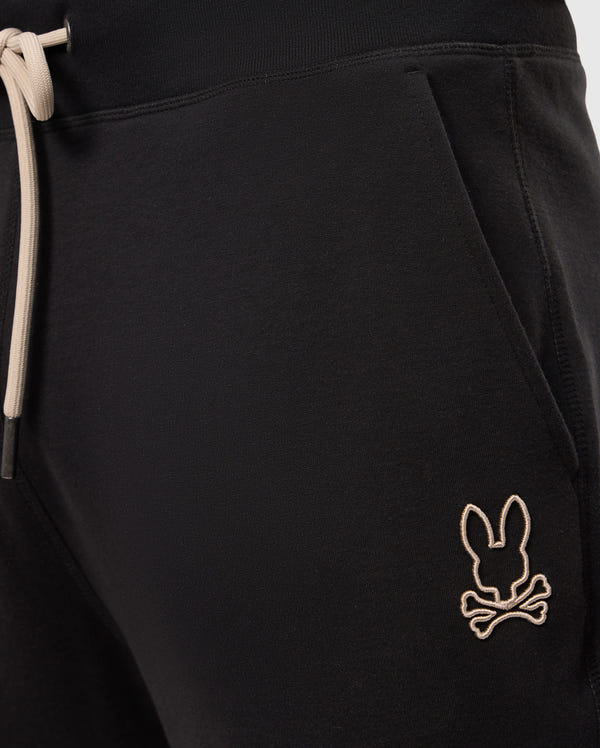Close-up of the MENS JAMES PREMIUM SWEATPANT - B6P791D200 by Psycho Bunny, designed with a beige drawstring against the black cotton-blend fabric. A small embroidered logo featuring a bunny head above two crossed bones on the front pocket adds a stylish touch to these premium sweatpants without compromising comfort.