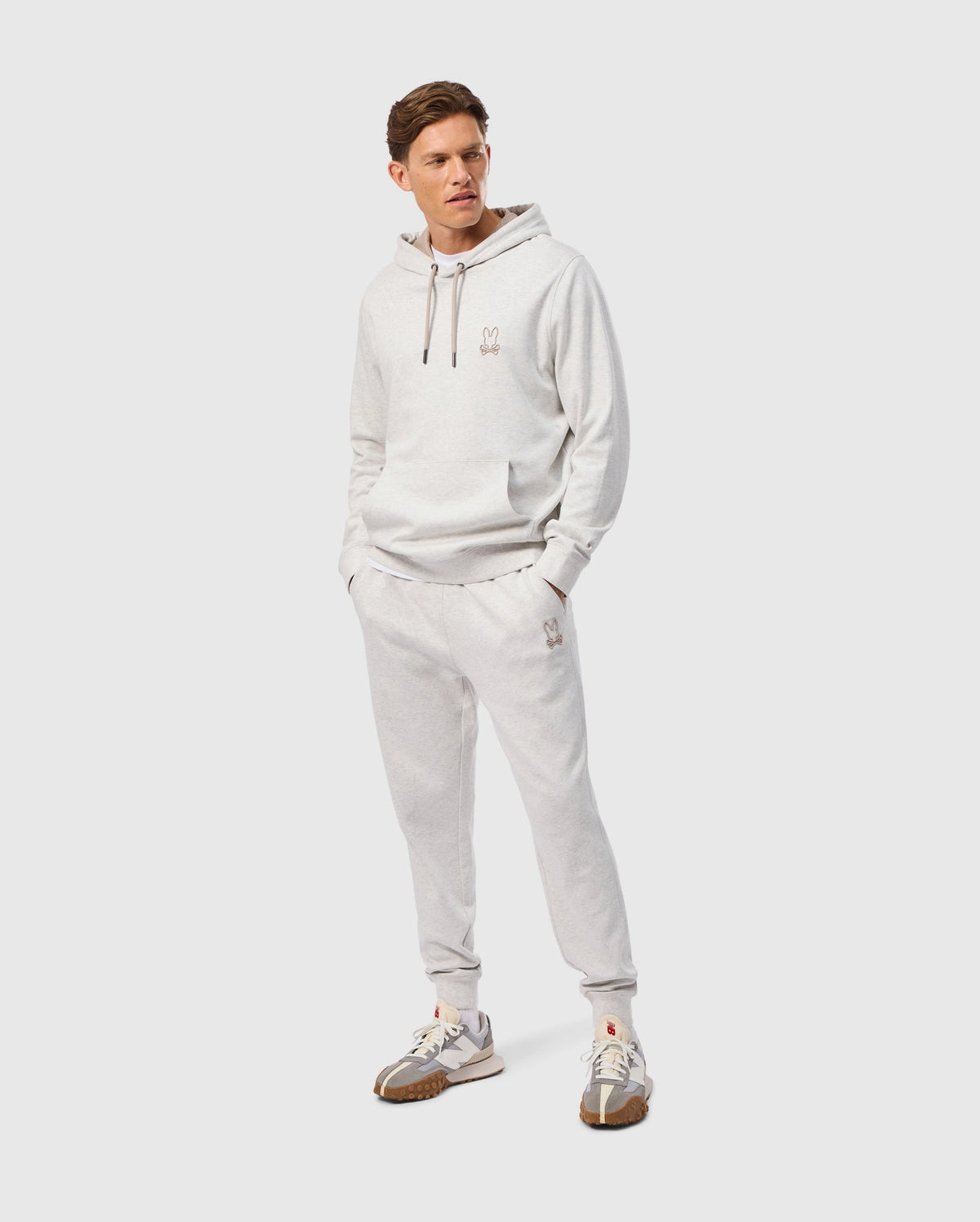 A person wearing a light gray hoodie and matching Psycho Bunny MENS JAMES PREMIUM SWEATPANT - B6P791D200 stands against a plain white background. The premium bottoms feature small logo embroidery on the leg, adding a subtle branding touch. They are also wearing white sneakers with brown accents, completing the casual yet stylish look.