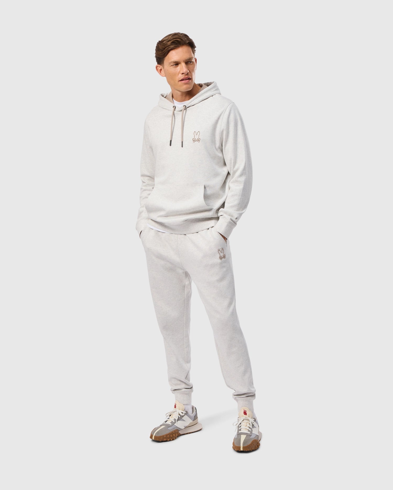 A person wearing a light gray hoodie and matching Psycho Bunny MENS JAMES PREMIUM SWEATPANT - B6P791D200 stands against a plain white background. The premium bottoms feature small logo embroidery on the leg, adding a subtle branding touch. They are also wearing white sneakers with brown accents, completing the casual yet stylish look.