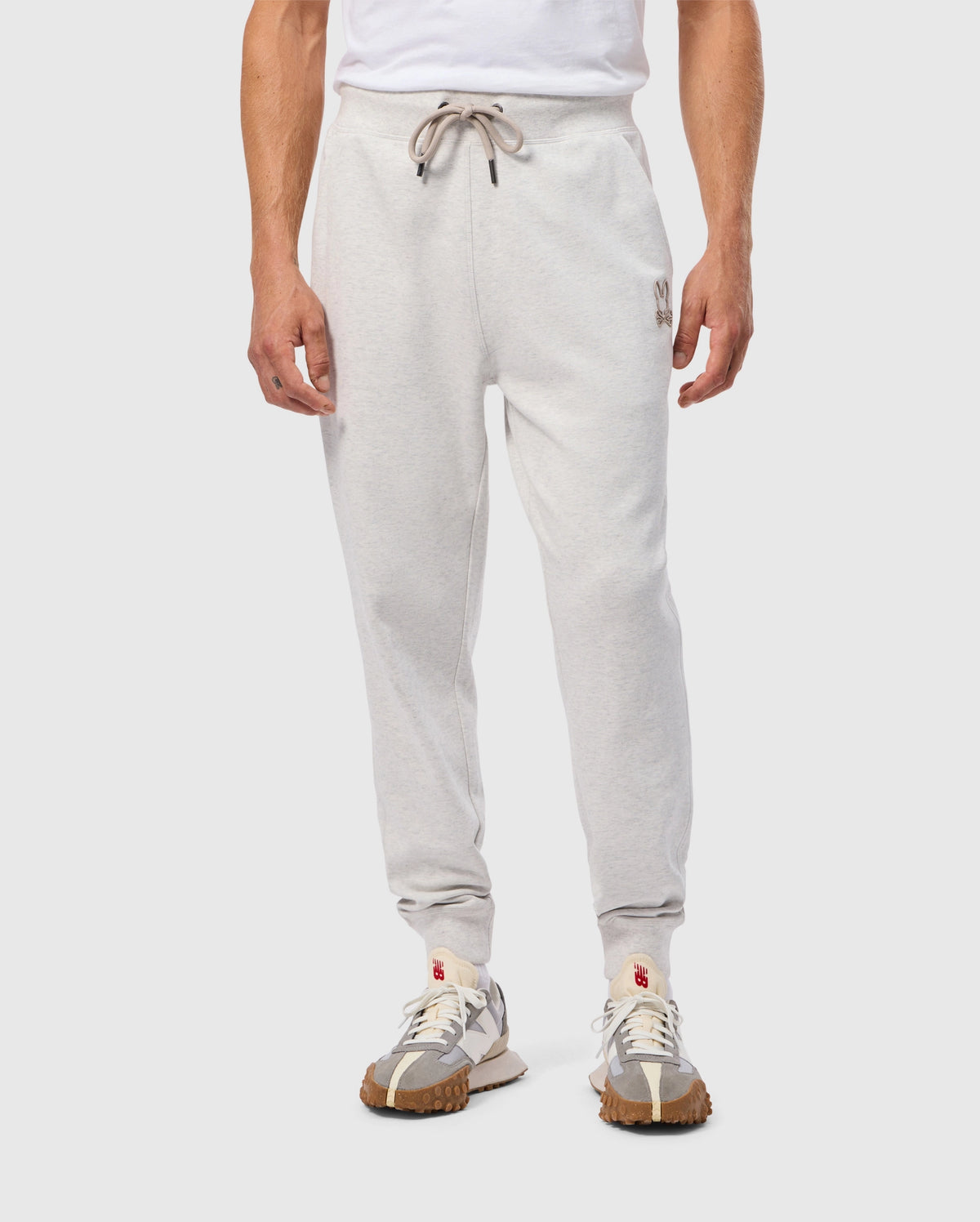 A person wearing the Psycho Bunny MENS JAMES PREMIUM SWEATPANT - B6P791D200, featuring light grey cotton-blend joggers with a drawstring waist and a small emblem on the left thigh, paired with a white T-shirt. They are also sporting white sneakers with brown textured soles.