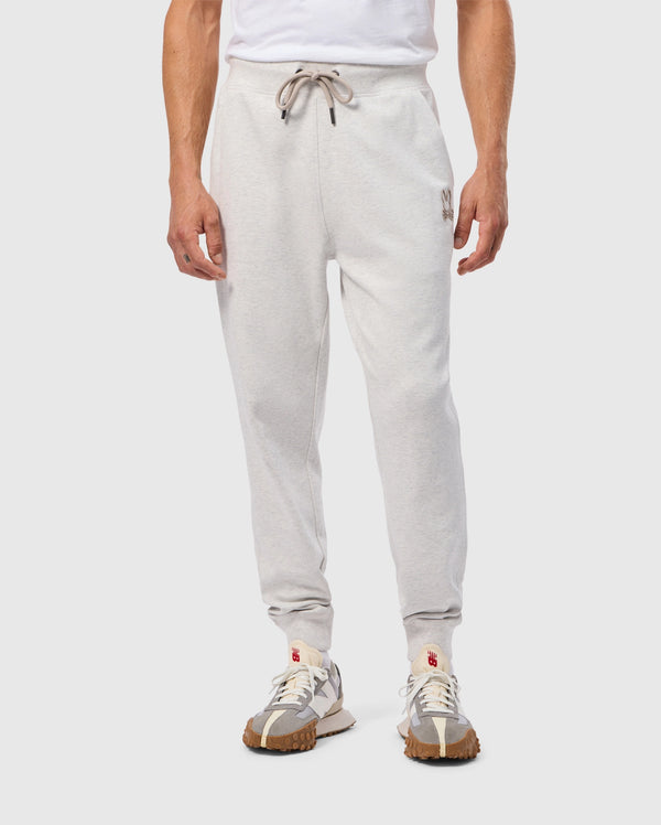A person wearing the Psycho Bunny MENS JAMES PREMIUM SWEATPANT - B6P791D200, featuring light grey cotton-blend joggers with a drawstring waist and a small emblem on the left thigh, paired with a white T-shirt. They are also sporting white sneakers with brown textured soles.