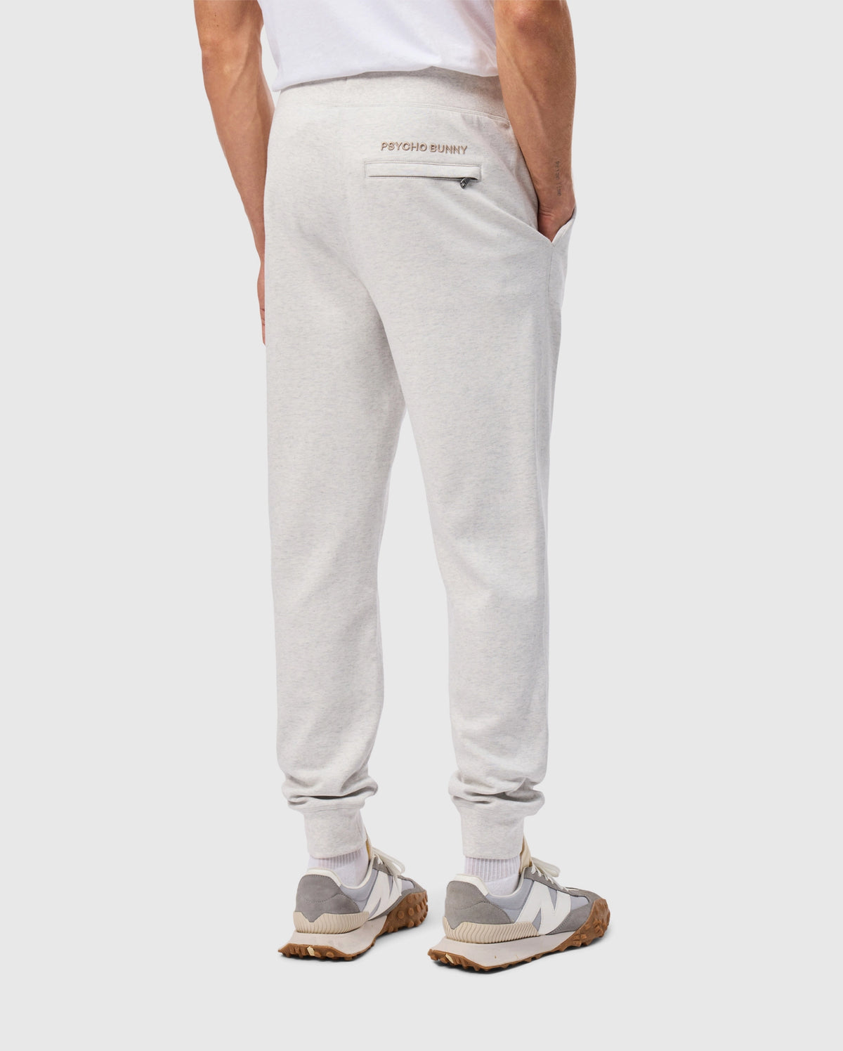 A person wearing light gray, cotton-blend MENS JAMES PREMIUM SWEATPANT from Psycho Bunny and a white T-shirt stands with hands in pockets. Their premium bottoms feature "Psycho Bunny" written above the back pocket, paired with white and gray sneakers.