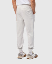 A person wearing light gray, cotton-blend MENS JAMES PREMIUM SWEATPANT from Psycho Bunny and a white T-shirt stands with hands in pockets. Their premium bottoms feature "Psycho Bunny" written above the back pocket, paired with white and gray sneakers.