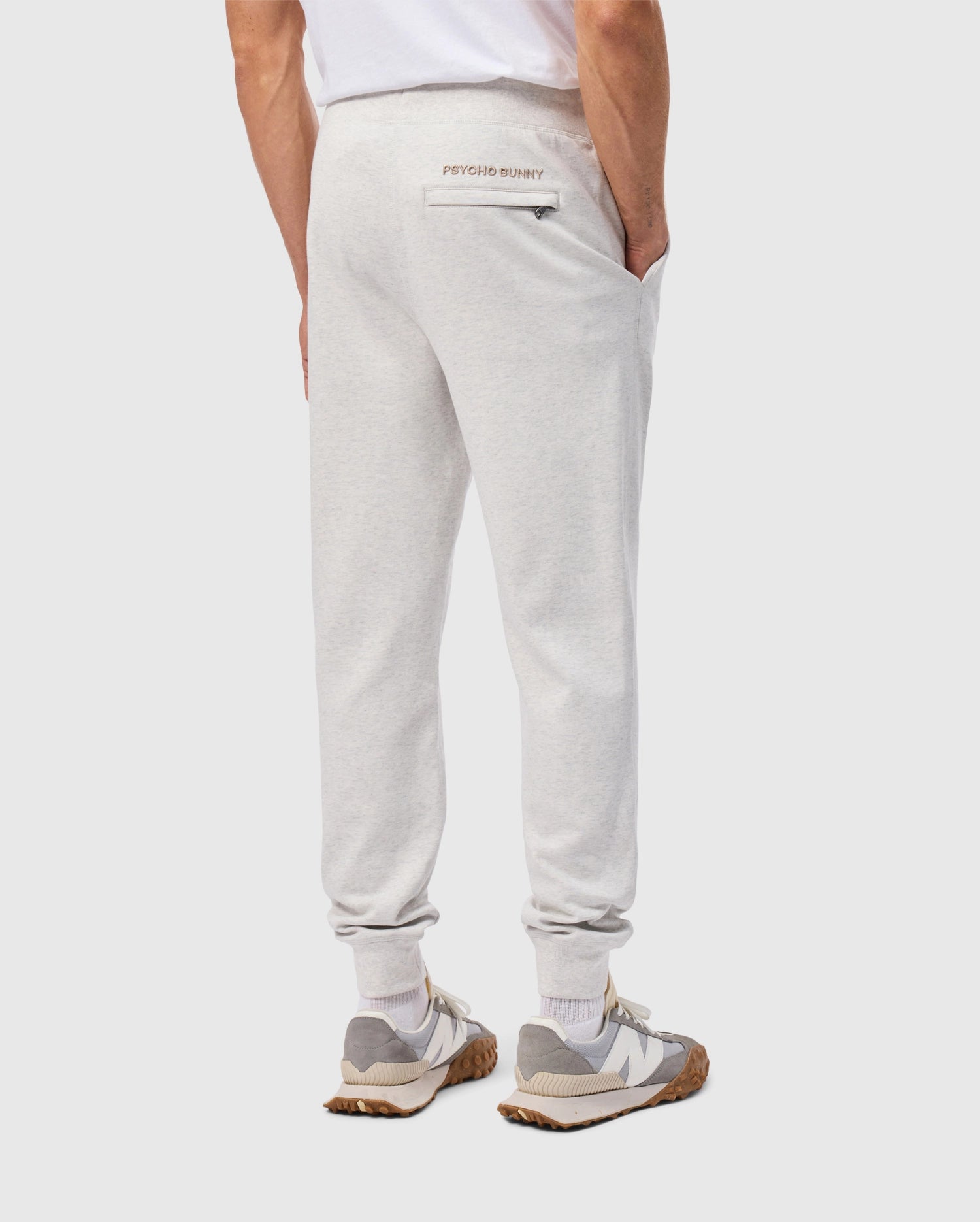 A person wearing light gray, cotton-blend MENS JAMES PREMIUM SWEATPANT from Psycho Bunny and a white T-shirt stands with hands in pockets. Their premium bottoms feature "Psycho Bunny" written above the back pocket, paired with white and gray sneakers.