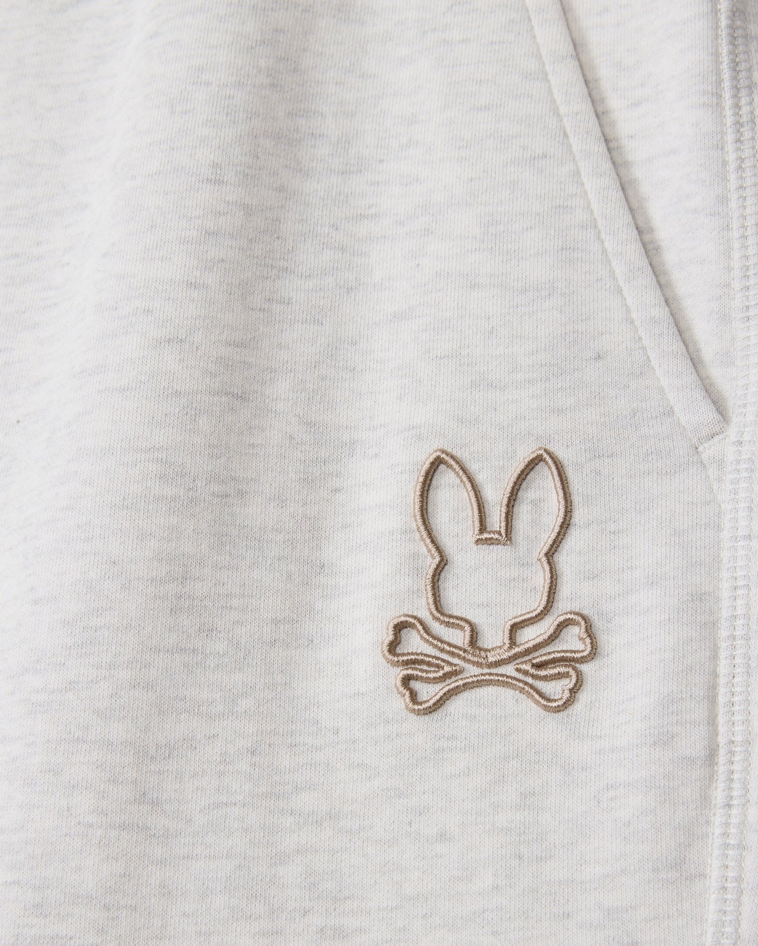 A close-up of the light gray fabric on the Psycho Bunny MENS JAMES PREMIUM SWEATPANT - B6P791D200 highlights a rabbit ears and crossbones logo embroidered near the seam. The logo in light beige offers a distinctive touch to these premium sweatpants, crafted from a soft cotton-blend.