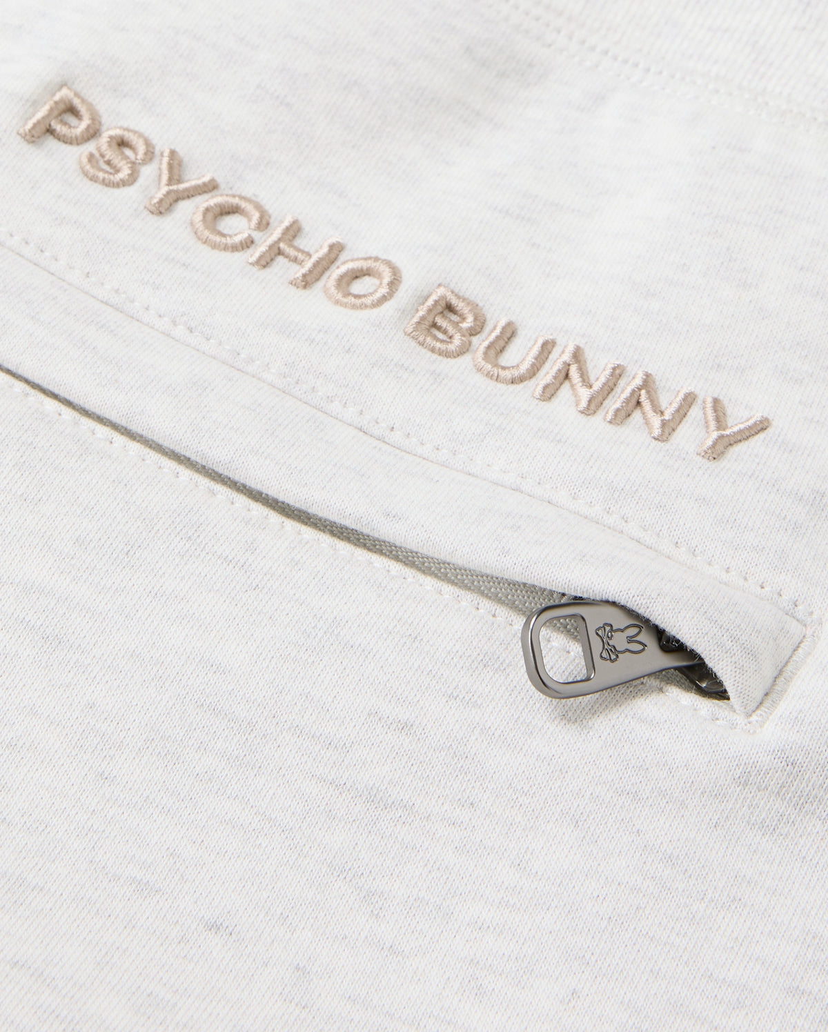 A detailed view of the cotton-blend fabric showcases "Psycho Bunny" elegantly embroidered in beige. Situated below is a small zipper adorned with a bunny icon on the pull tab, injecting a playful element into these MENS JAMES PREMIUM SWEATPANT - B6P791D200, echoing the classic design of James sweatpants by Psycho Bunny.