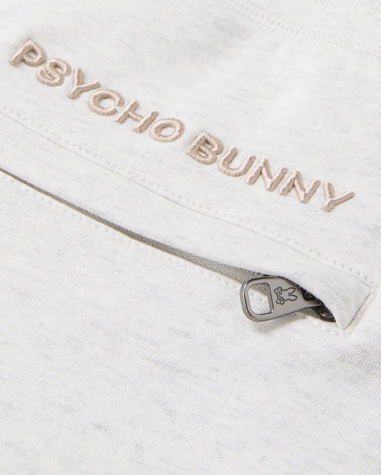 A detailed view of the cotton-blend fabric showcases "Psycho Bunny" elegantly embroidered in beige. Situated below is a small zipper adorned with a bunny icon on the pull tab, injecting a playful element into these MENS JAMES PREMIUM SWEATPANT - B6P791D200, echoing the classic design of James sweatpants by Psycho Bunny.
