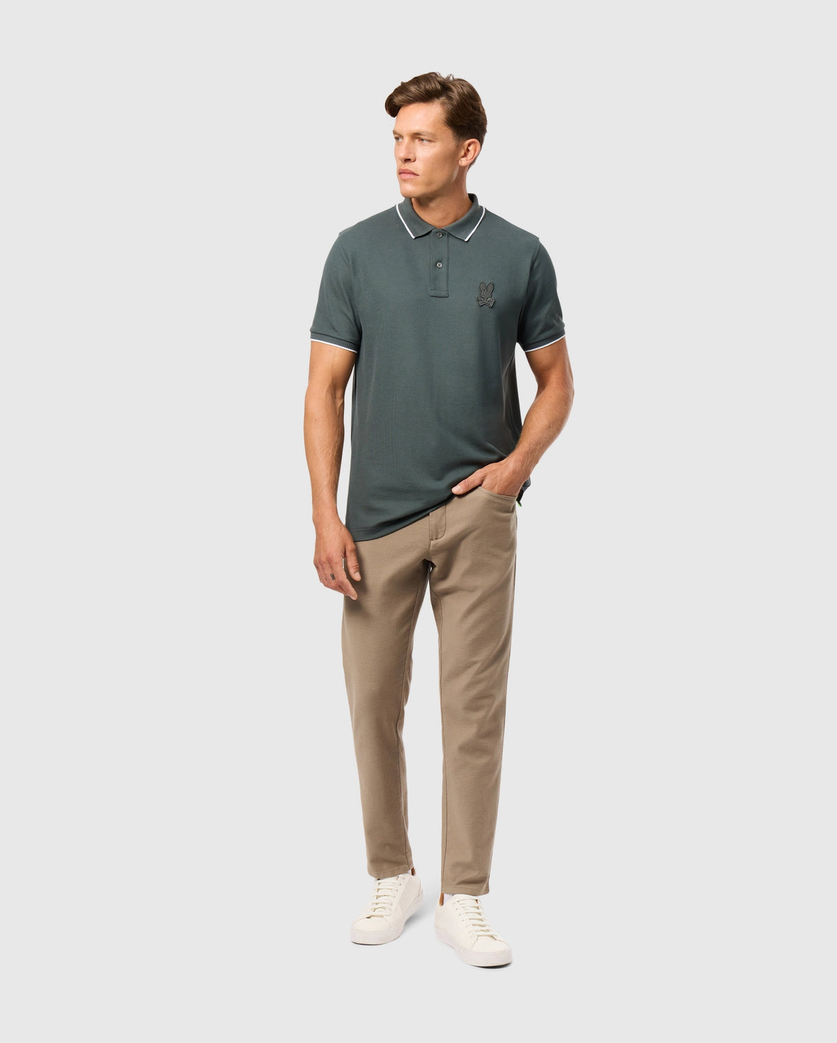 A person wearing a dark green polo shirt and the Psycho Bunny MENS HAYS TWILL PANT in beige stands against a plain background. They have their right hand in the pocket of the 5-pocket pants and are wearing white sneakers.