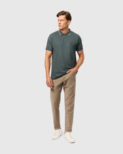 A person wearing a dark green polo shirt and the Psycho Bunny MENS HAYS TWILL PANT in beige stands against a plain background. They have their right hand in the pocket of the 5-pocket pants and are wearing white sneakers.