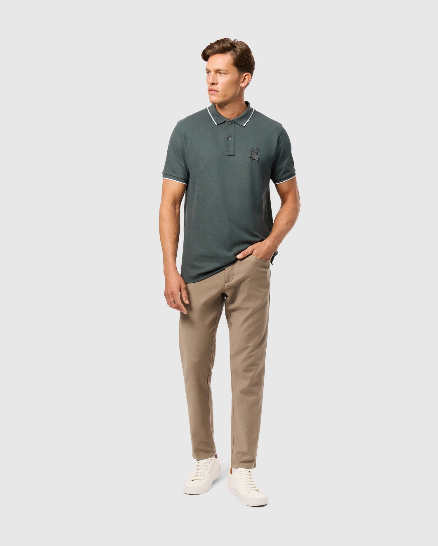 A person wearing a dark green polo shirt and the Psycho Bunny MENS HAYS TWILL PANT in beige stands against a plain background. They have their right hand in the pocket of the 5-pocket pants and are wearing white sneakers.