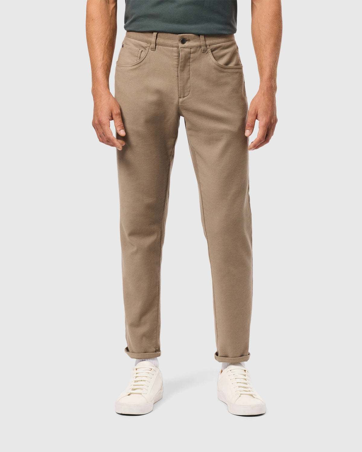 A person stands against a neutral background wearing Psycho Bunny’s MENS HAYS TWILL PANT - B6P894D200, which are cotton-blend with a straight fit and five-pocket design, in tan, slightly rolled at the cuffs. They pair these pants with white sneakers and a dark green top, keeping their hands at their sides.