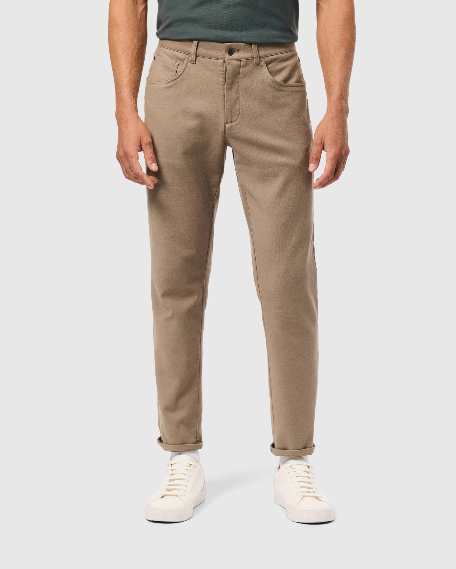 A person stands against a neutral background wearing Psycho Bunny’s MENS HAYS TWILL PANT - B6P894D200, which are cotton-blend with a straight fit and five-pocket design, in tan, slightly rolled at the cuffs. They pair these pants with white sneakers and a dark green top, keeping their hands at their sides.