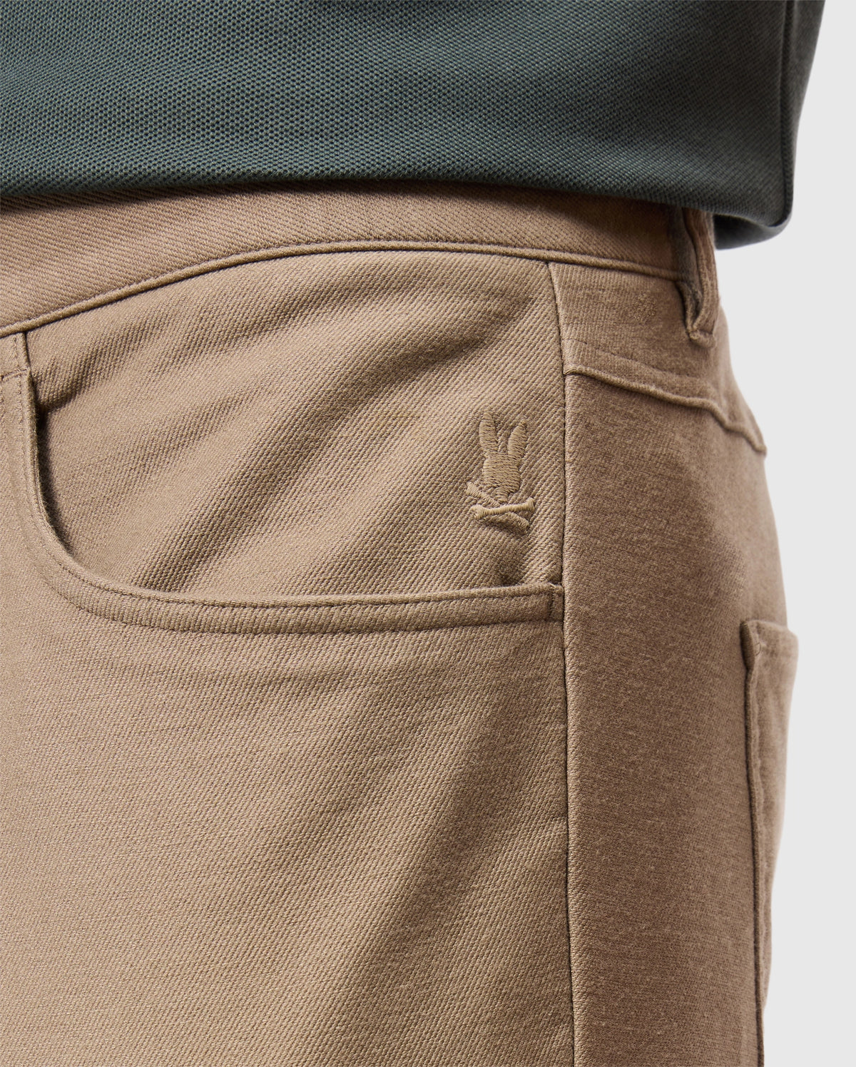Close-up of a person's waist wearing the MENS HAYS TWILL PANT - B6P894D200 by Psycho Bunny, featuring a tan cotton-blend twill fabric with a five-pocket design and a small embroidered bunny logo on the front pocket. The person is also wearing a dark green top.