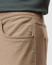 Close-up of a person's waist wearing the MENS HAYS TWILL PANT - B6P894D200 by Psycho Bunny, featuring a tan cotton-blend twill fabric with a five-pocket design and a small embroidered bunny logo on the front pocket. The person is also wearing a dark green top.