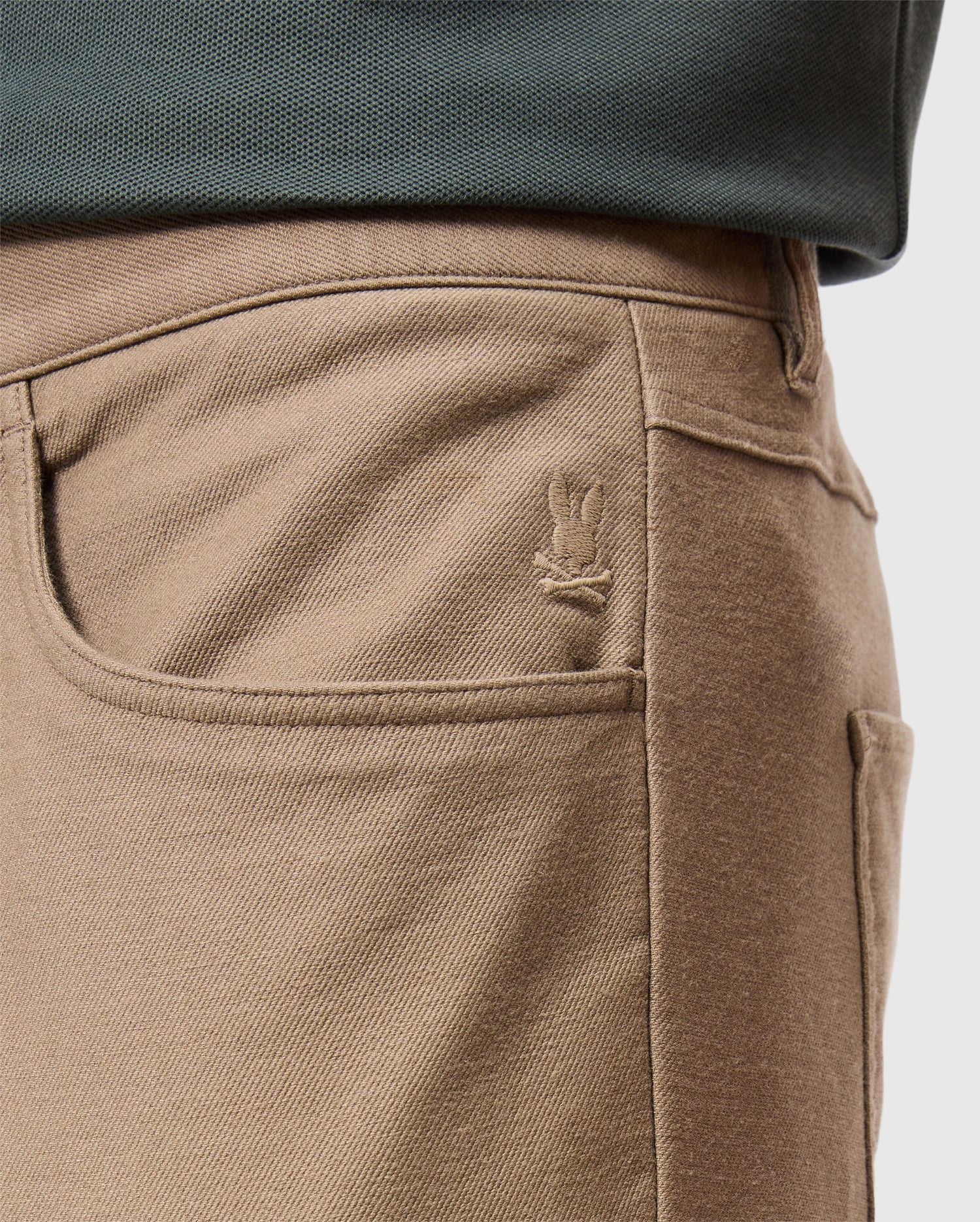 Close-up of a person's waist wearing the MENS HAYS TWILL PANT - B6P894D200 by Psycho Bunny, featuring a tan cotton-blend twill fabric with a five-pocket design and a small embroidered bunny logo on the front pocket. The person is also wearing a dark green top.