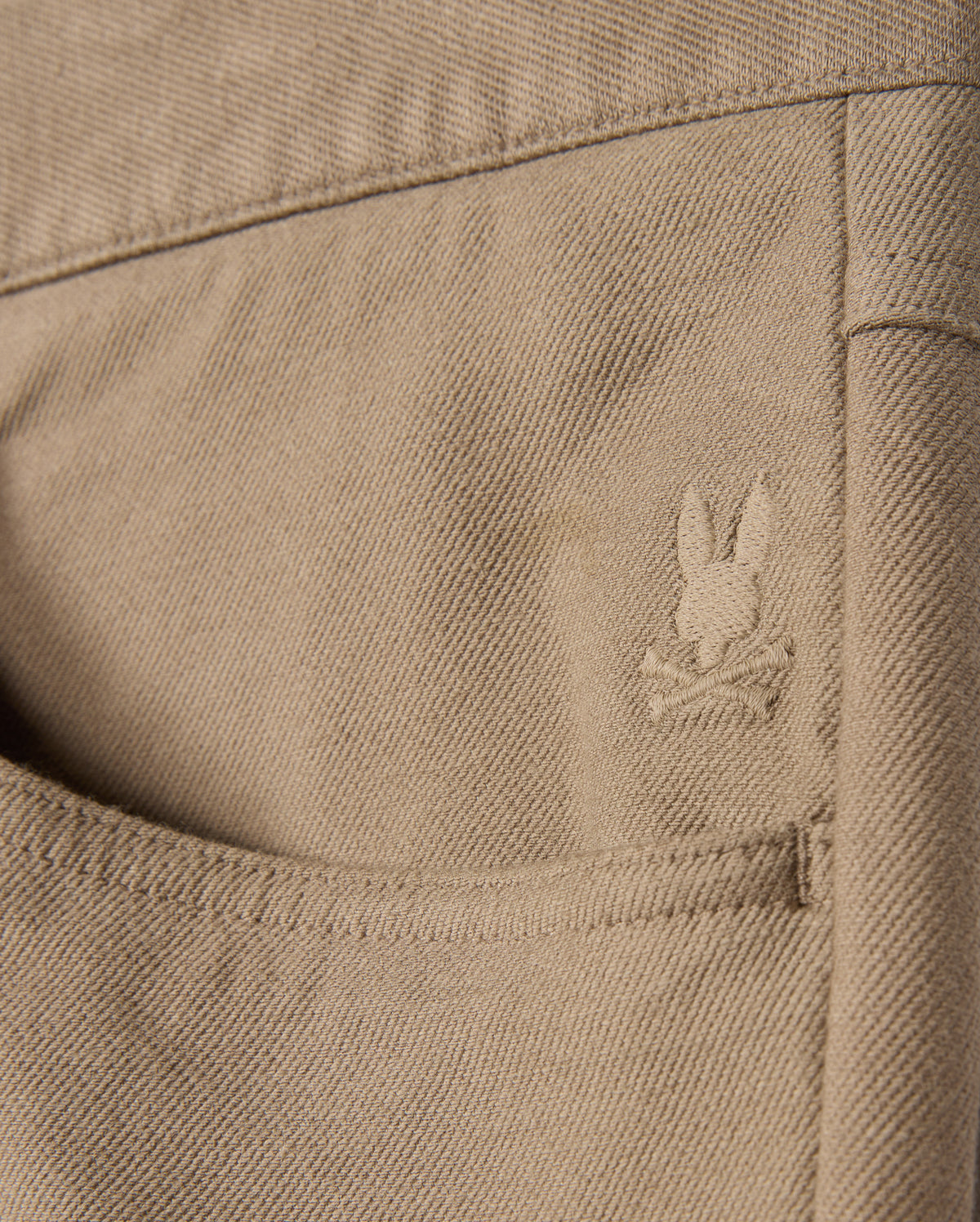The MENS HAYS TWILL PANT - B6P894D200 by Psycho Bunny features a close-up of a khaki cotton-blend twill pocket on its five-pocket design, adorned with an embroidered rabbit head logo and crossbones. The fabric's texture and stitching details are enhanced by its innovative four-way stretch design.