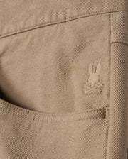 The MENS HAYS TWILL PANT - B6P894D200 by Psycho Bunny features a close-up of a khaki cotton-blend twill pocket on its five-pocket design, adorned with an embroidered rabbit head logo and crossbones. The fabric's texture and stitching details are enhanced by its innovative four-way stretch design.