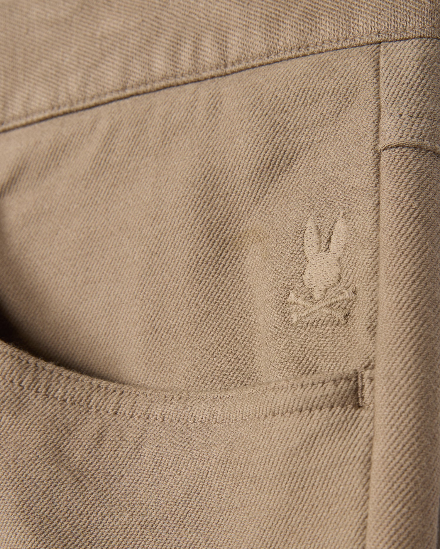 The MENS HAYS TWILL PANT - B6P894D200 by Psycho Bunny features a close-up of a khaki cotton-blend twill pocket on its five-pocket design, adorned with an embroidered rabbit head logo and crossbones. The fabric's texture and stitching details are enhanced by its innovative four-way stretch design.