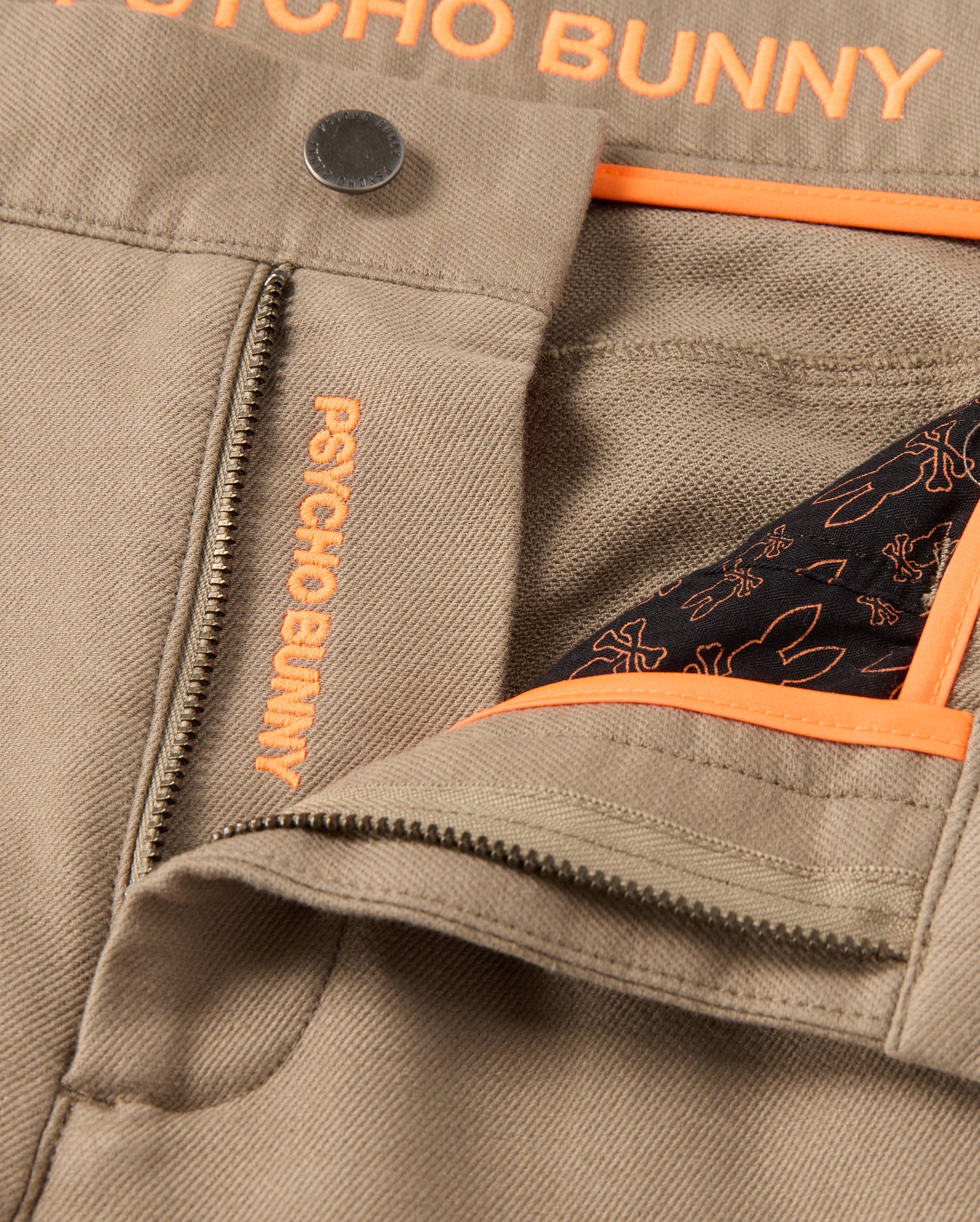Zoomed image of the Mens Hays Twill Pant - B6P894D200 in a khaki cotton-blend twill, highlighted by a zipper and button closure. The signature orange "Psycho Bunny" branding is prominently showcased on the inner lining and near the zipper, complemented by a contrasting pattern on the pocket lining. This pant is crafted with five pockets and four-way stretch material for enhanced comfort.