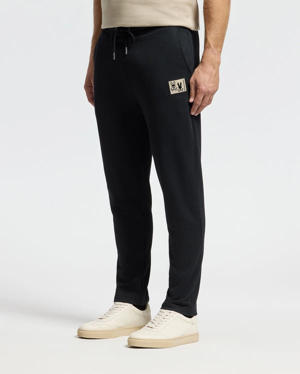 A person wearing sleek black Psycho Bunny Monroe Twill Patch Fleece Sweatpants, featuring a small logo patch on the upper thigh, pairs them with a beige shirt and white sneakers while standing against a plain white background.