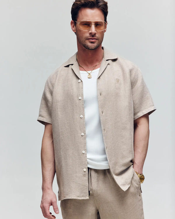 A man stands against a plain background wearing the Psycho Bunny MENS WINDCREST LINEN BLEND SHORT SLEEVE SHIRT (B6Q148C200) in beige over a white tank, paired with beige pants, a gold watch, necklaces, and brown-tinted sunglasses.