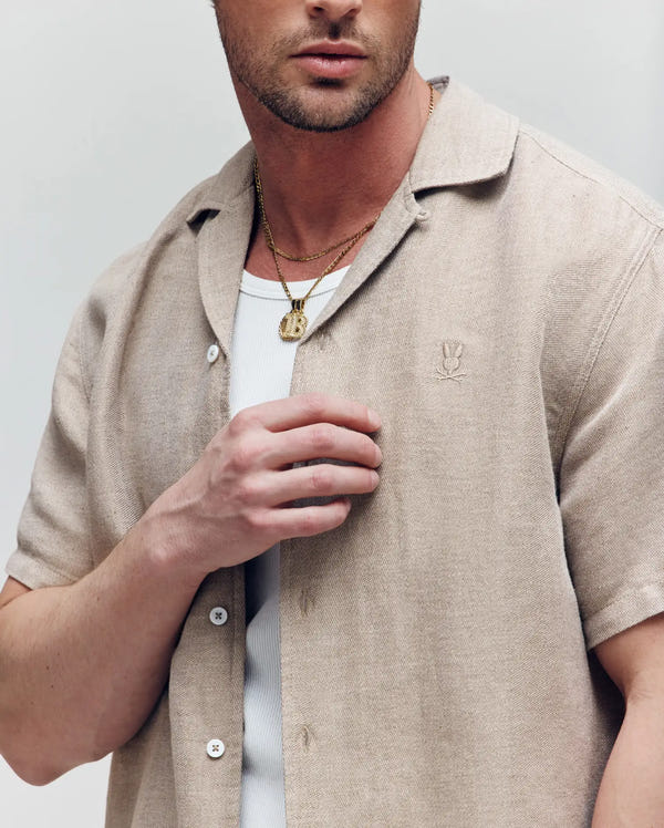 A man stands against a light background wearing the Psycho Bunny MENS WINDCREST LINEN BLEND SHORT SLEEVE SHIRT (B6Q148C200) in beige over a white ribbed tee, layered with gold necklaces. His face is partially out of frame.