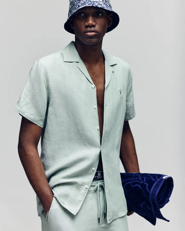 A man in a light green Psycho Bunny MENS WINDCREST LINEN BLEND SHORT SLEEVE SHIRT (B6Q148C200), matching pants, and a patterned bucket hat holds a dark blue towel under his arm against a plain, light background.