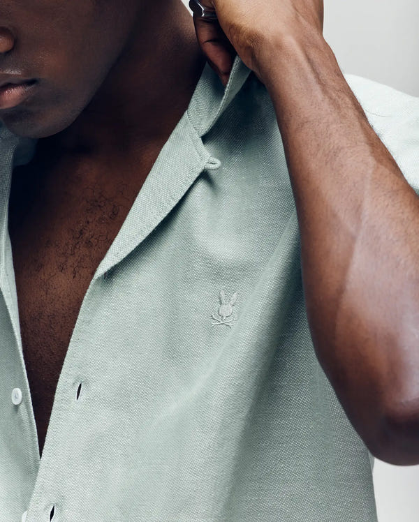 A close-up shows a person adjusting the collar of the Psycho Bunny Mens Windcrest Linen Blend Short Sleeve Shirt (B6Q148C200), featuring light green linen-cotton fabric, an embroidered chest logo, and Mother-of-Pearl buttons. Lower face and torso visible.