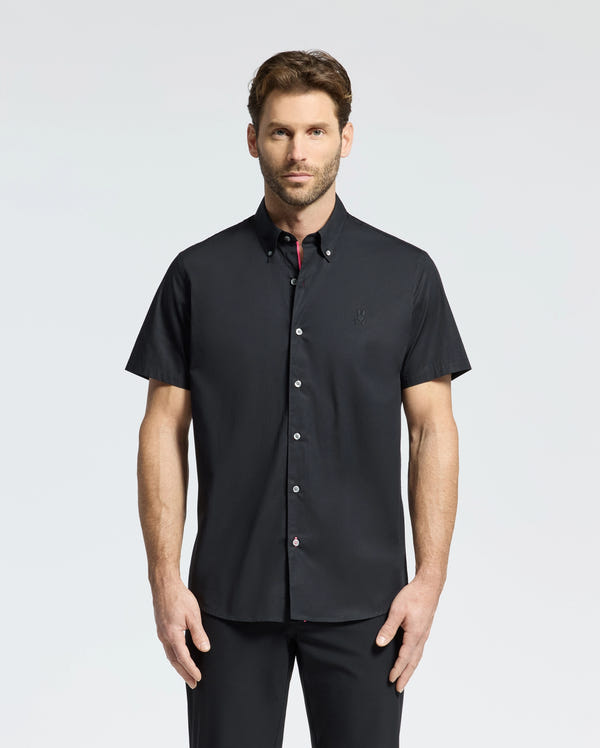 A man stands against a plain background wearing a Psycho Bunny MENS GABRIEL SHORT SLEEVE SHIRT in black, paired with matching pants. Crafted from premium Pima cotton, the ensemble complements his direct gaze and neutral expression.