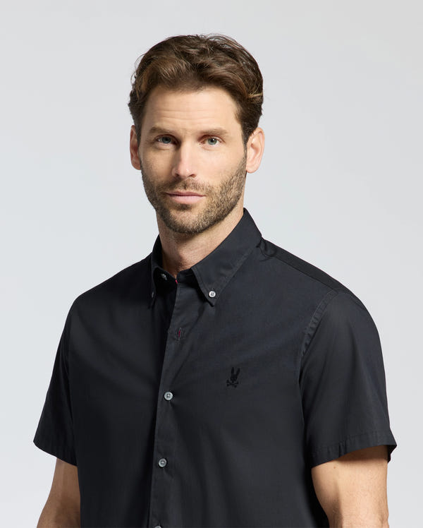 A man with short brown hair and a beard wears a black Psycho Bunny MENS GABRIEL SHORT SLEEVE SHIRT, B6Q351F200, made of woven stretch Pima cotton. He stands against a plain light background, gazing at the camera with a neutral expression.