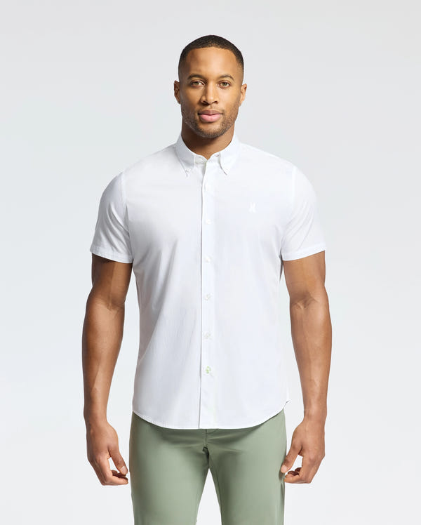 Against a light background, a man wears Psycho Bunny's MENS GABRIEL SHORT SLEEVE SHIRT in white, made from Pima cotton, with light green pants. An embroidered bunny adds charm to his outfit as he gazes at the camera with a neutral expression.