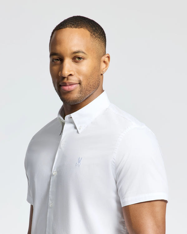 A man in a white MENS GABRIEL SHORT SLEEVE SHIRT (B6Q351F200) from Psycho Bunny, crafted from Pima cotton with an embroidered bunny, stands against a plain background and smiles at the camera.