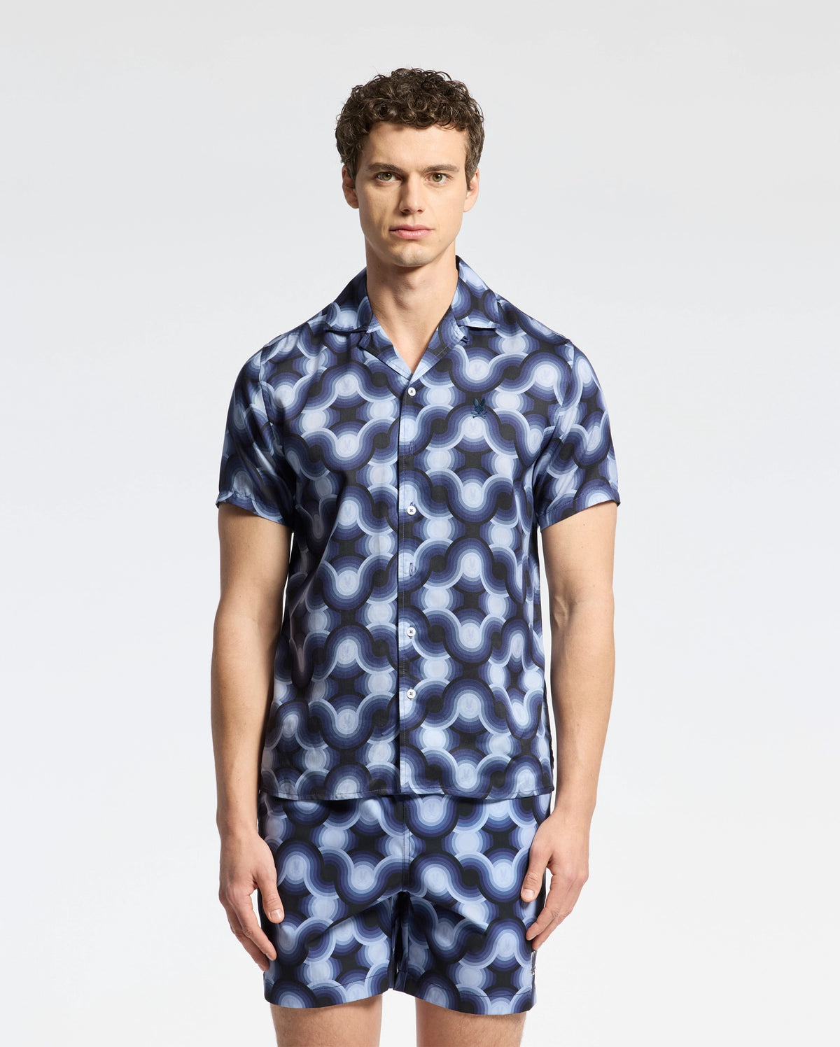 A man with curly hair wears the MENS PAROS TENCEL SHIRT from Psycho Bunny and matching shorts, both featuring a blue and black geometric pattern. He stands against a white background, showcasing effortless style.