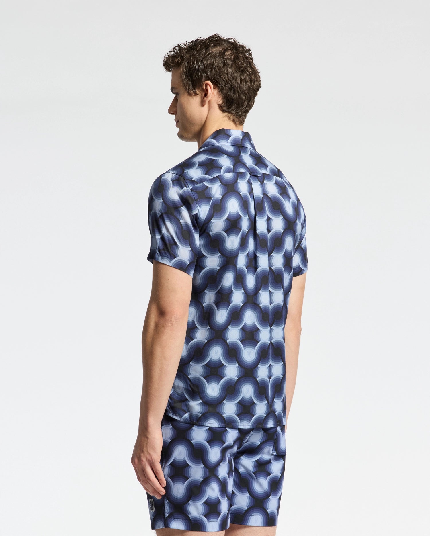 A person stands sideways wearing Psycho Bunny's MENS PAROS TENCEL SHIRT - B6Q460F200, a matching blue geometric-patterned shirt and shorts set with a wavy design, embodying the essence of a good-time shirt against a light-colored plain background.