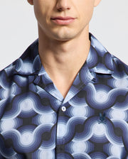 A person wears the Psycho Bunny Men's Paros Tencel Shirt, B6Q460F200. The blue and white circular wave-patterned, collared shirt is made of woven Tencel and sports a small embroidered chest logo. Only the lower half of their face and upper torso are shown.