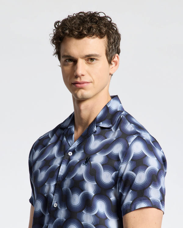 Wearing a Psycho Bunny MENS PAROS TENCEL SHIRT - B6Q460F200 with blue interlocking circular patterns, the curly-haired person stands confidently against a plain background, ready to embrace the day.