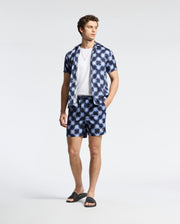 A man wears the Psycho Bunny MENS PAROS TENCEL SHIRT paired with matching blue geometric-patterned shorts, a white T-shirt underneath, and black sandals. His hand rests in his pocket, against a plain light background enhancing the chill vibe.