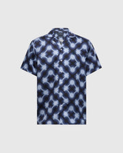 The MENS PAROS TENCEL SHIRT - B6Q460F200 by Psycho Bunny is a short-sleeve button-up styled in a blue and grey retro wave pattern on a light background, crafted from woven Tencel with a classic collar and matching buttons.