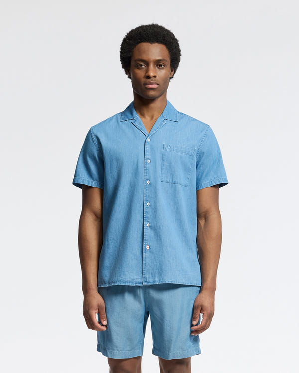 A person stands against a plain background wearing Psycho Bunny's MENS KERVIN LIGHTWEIGHT DENIM WOVEN SHIRT (B6Q467F200), a casual light blue, short-sleeved button-up shirt with subtle chest embroidery and matching shorts. The lightweight material is perfect for warm weather.