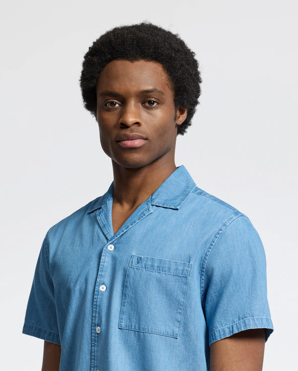 A person with short, curly hair wearing the Psycho Bunny MENS KERVIN LIGHTWEIGHT DENIM WOVEN SHIRT - B6Q467F200 looks directly at the camera against a plain white background.