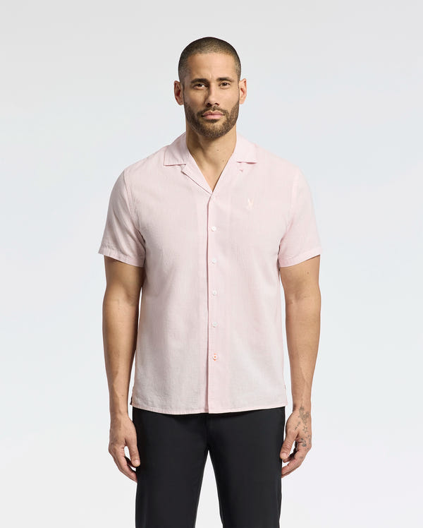 A bearded man with short hair stands against a plain backdrop, wearing Psycho Bunny's MENS COBAR WOVEN SHIRT - B6Q522F200, a light pink linen blend short-sleeve shirt with subtle embroidery along the collar, paired with black pants. Tattoos are displayed on his left hand.