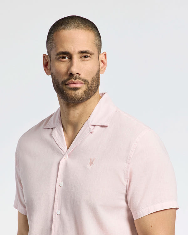A bearded person is wearing the Psycho Bunny MENS COBAR WOVEN SHIRT - B6Q522F200, a light pink, short-sleeved linen blend with delicate chest embroidery, standing against a plain light background.
