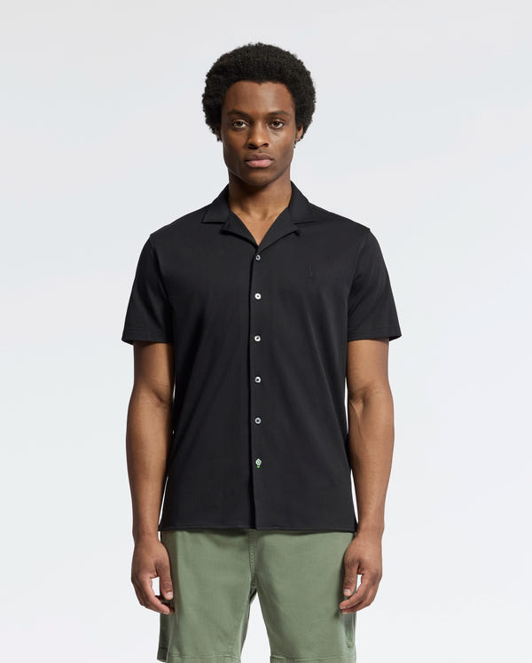 A person wearing Psycho Bunny's MENS BRYAN CAMP COLLAR SHORT SLEEVE SHIRT, made of moisture-wicking Pima cotton and featuring a button-up design in black, stands with green shorts against a plain background. Their expression is neutral, and their hands rest by their sides.