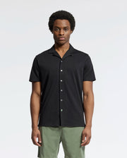 A person wearing Psycho Bunny's MENS BRYAN CAMP COLLAR SHORT SLEEVE SHIRT, made of moisture-wicking Pima cotton and featuring a button-up design in black, stands with green shorts against a plain background. Their expression is neutral, and their hands rest by their sides.