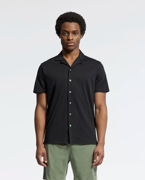 A person wearing Psycho Bunny's MENS BRYAN CAMP COLLAR SHORT SLEEVE SHIRT, made of moisture-wicking Pima cotton and featuring a button-up design in black, stands with green shorts against a plain background. Their expression is neutral, and their hands rest by their sides.