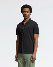 A person stands against a plain background wearing Psycho Bunny's Mens Bryan Camp Collar Short Sleeve Shirt (B6Q581F200) in black, paired with green pants. Made from Pima cotton, the shirt ensures comfort. They face forward with a neutral expression.