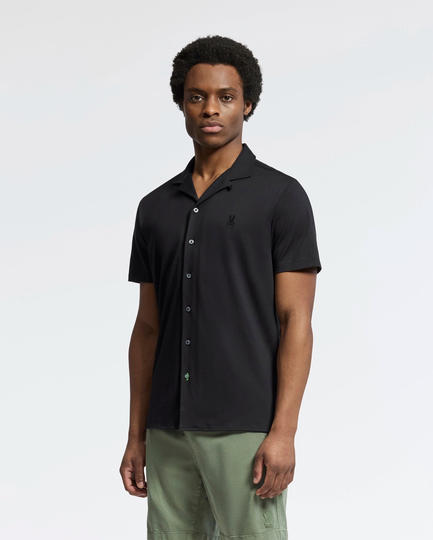 A person stands against a plain background wearing Psycho Bunny's Mens Bryan Camp Collar Short Sleeve Shirt (B6Q581F200) in black, paired with green pants. Made from Pima cotton, the shirt ensures comfort. They face forward with a neutral expression.