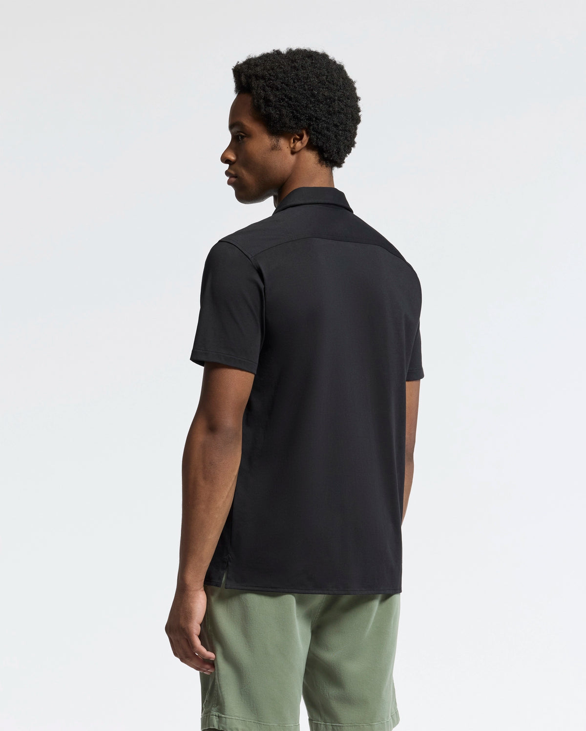 A person with short curly hair, slightly turned away, wears the Psycho Bunny MENS BRYAN CAMP COLLAR SHORT SLEEVE SHIRT in black and olive green shorts. The light background emphasizes their outfit's moisture-wicking fabric.